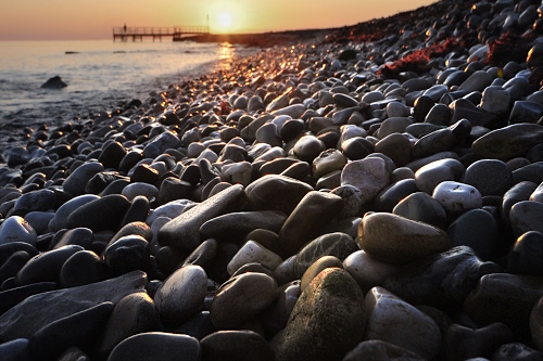 Immagine con tramonto, mare, sassi, sole, pontile, spiaggia, acqua, riflessi, pietre, ciottoli, rosso, grigio, luce, prospettiva, controluce, cielo