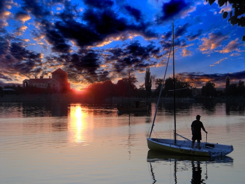 Immagine con barca, nuvole, cielo, acqua, tramonto, sole, controluce, riflesso, mare, lago, pescatore, riflessi, azzurro, case, alberi, persona