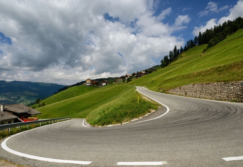 Immagine con strada, curva, verde, nuvole, tornante, cielo, montagna, erba, prato, case