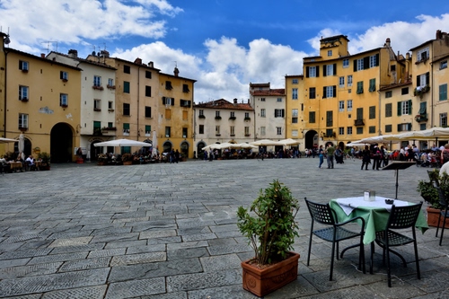 Immagine con piazza, ombrelloni, nuvole, sedie, case, cielo, finestre, archi, vaso, tavolini, piante, bar, tavoli, vasi, persone, palazzi
