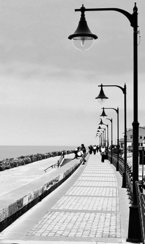 Immagine con lampioni, mare, bianconero, persone, lungomare, cielo, scogli, prospettiva, strada, scogliera, passeggiata, frangiflutti, casa, pali, case, passanti