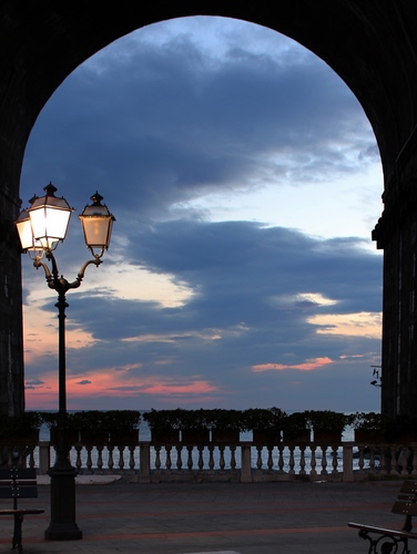Immagine con arco, nuvole, lampione, cielo, vasi, lampioni, balaustra, piante, tramonto, affaccio, sera, controluce, panchine, luci, piazza, fiori