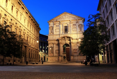 Immagine con chiesa, piazza, alberi, cielo, panchine, palazzi, statua, blu, finestre, monumento, luci, lampioni, persone, imbrunire