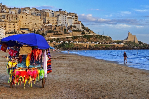 Immagine con mare, spiaggia, ombrellone, nuvole, cielo, sabbia, bancarella, case, venditore, paese, colori, costumi, borgo, castello, blu, riva, acqua, torre