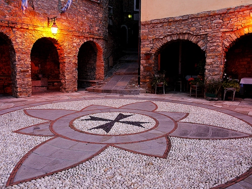 Immagine con croce, archi, sedie, luci, piazza, sassi, lampione, bandiere, pavimento, nero, portici, mosaico, notturno, stemma, malta, cerchi