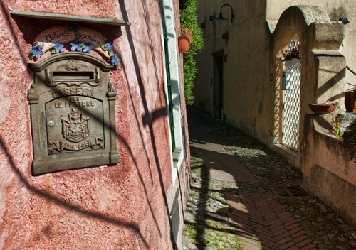 Immagine con vicolo, cassetta, cancello, posta, stradina, muro, lettere, vasi, ombre, verde, case, porta, buca, lampione, grata, finestra, pianta, rosso, fiori, via, strada, portone, arco, ombra, rosa