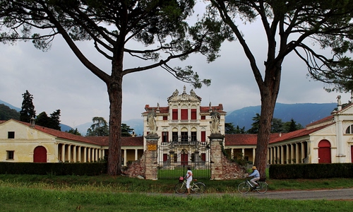 Immagine con alberi, cancello, villa, finestre, biciclette, cielo, persone, verde, colonne, portici, statue, pini, palazzo, porte, due, rami, erba, bici, tetto, archi, nuvole, bicicletta, ciclisti, edificio